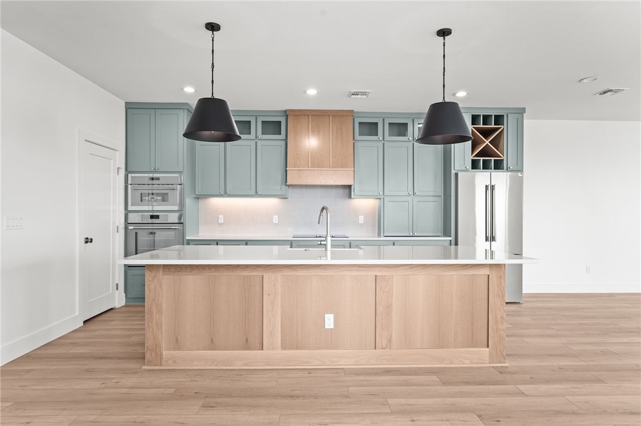 Kitchen at 8118 Beach Break with sage-green shaker cabinetry, a white oak waterfall island, a wood range hood, dark pendant lights and double wall ovens