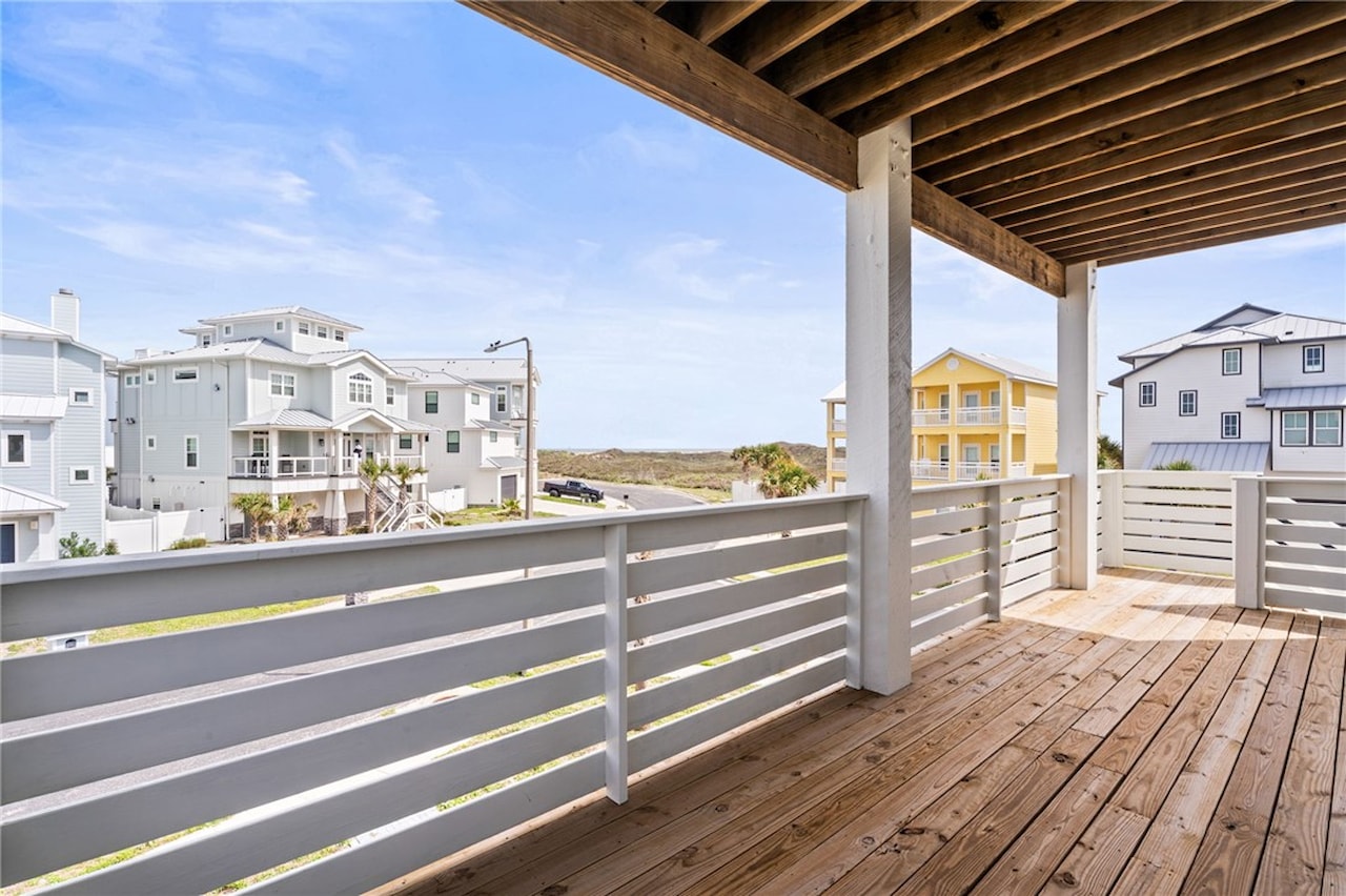 Covered second-floor balcony with white oak decking and horizontal white railing looking out over neighboring coastal homes toward open dune and sky