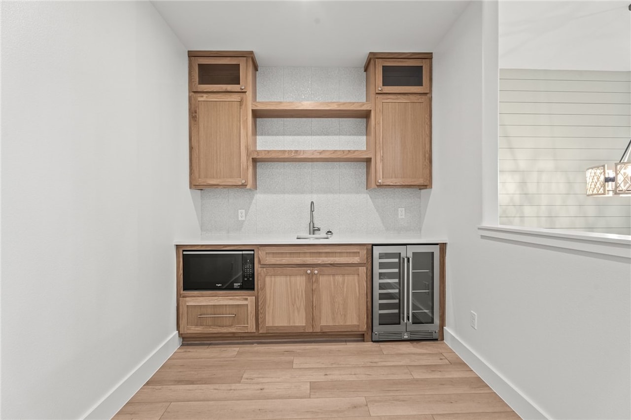 Wet bar and coffee station with light oak cabinetry, open shelving, a bar sink, a black microwave and a stainless wine cooler